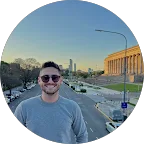 Homem sorridente de óculos escuros em uma avenida ao entardecer, com skyline urbano ao fundo.