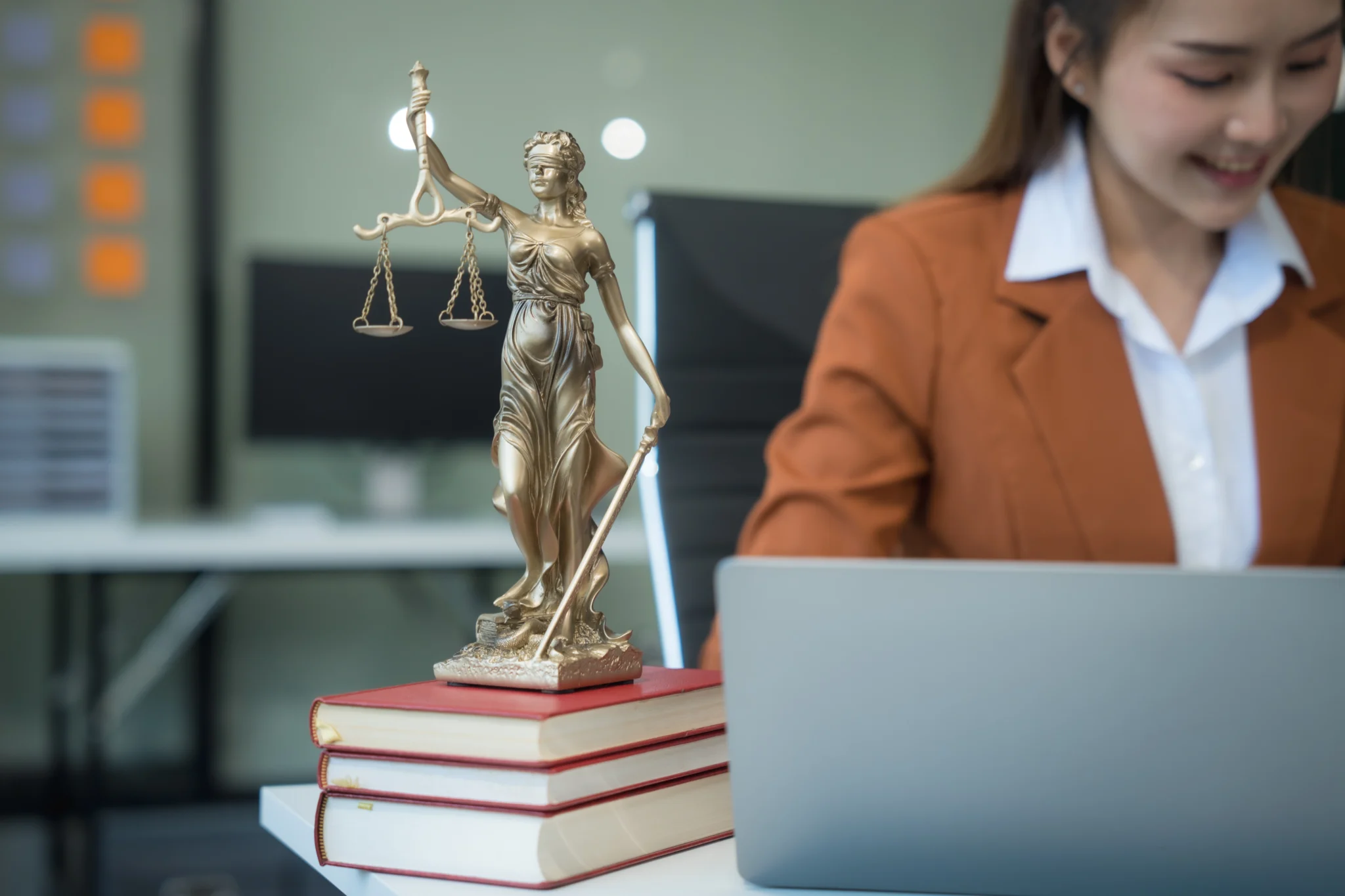 Estátua da deusa da justiça sobre pilha de livros jurídicos em primeiro plano, com advogada sorridente trabalhando no laptop ao fundo, simbolizando equilíbrio e estudo do direito.