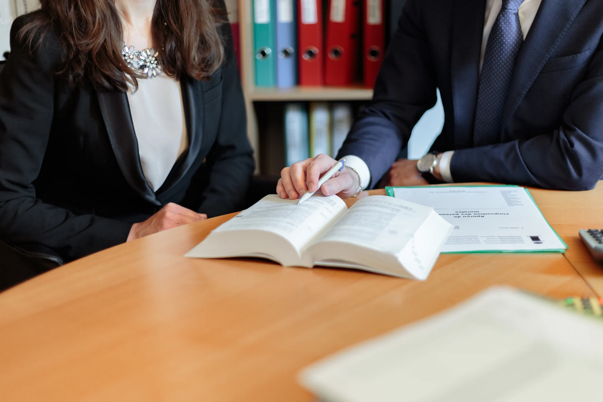 Advogados em trajes formais consultam livro jurídico aberto sobre a mesa, apontando um trecho com caneta enquanto analisam documentos do caso.