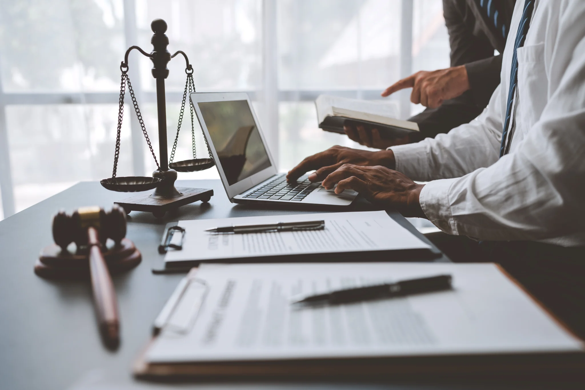 Advogados revisam documentos em laptop, com balança da justiça e martelo sobre a mesa, simbolizando análise jurídica e equilíbrio legal.