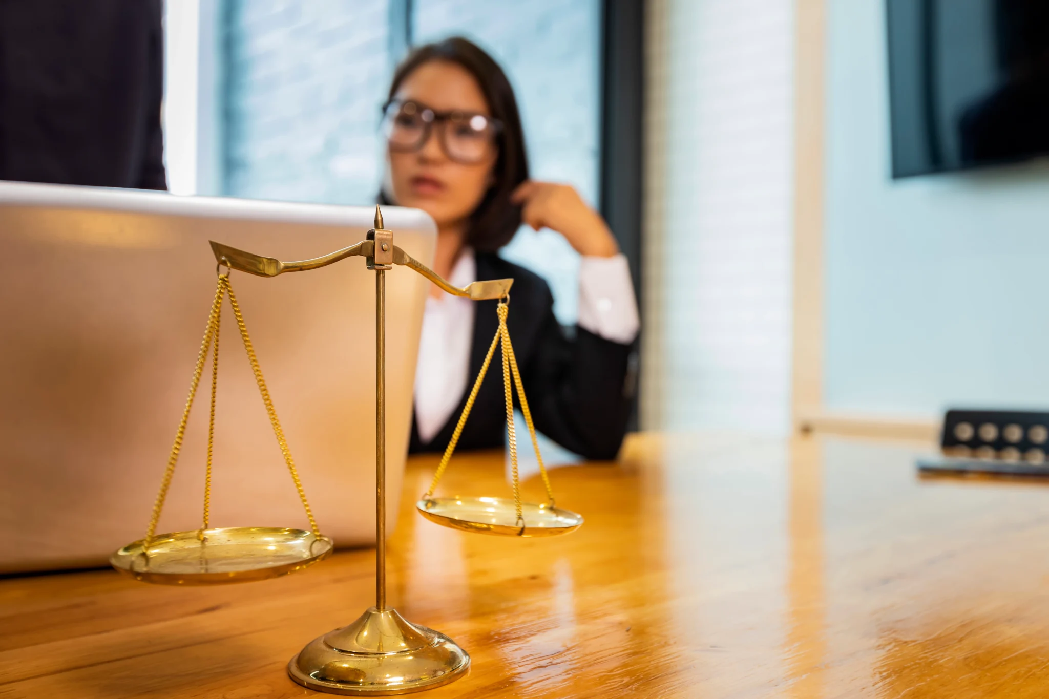 Balança dourada da justiça sobre mesa de madeira em primeiro plano, com advogada desfocada ao fundo trabalhando no notebook, representando equidade no direito.