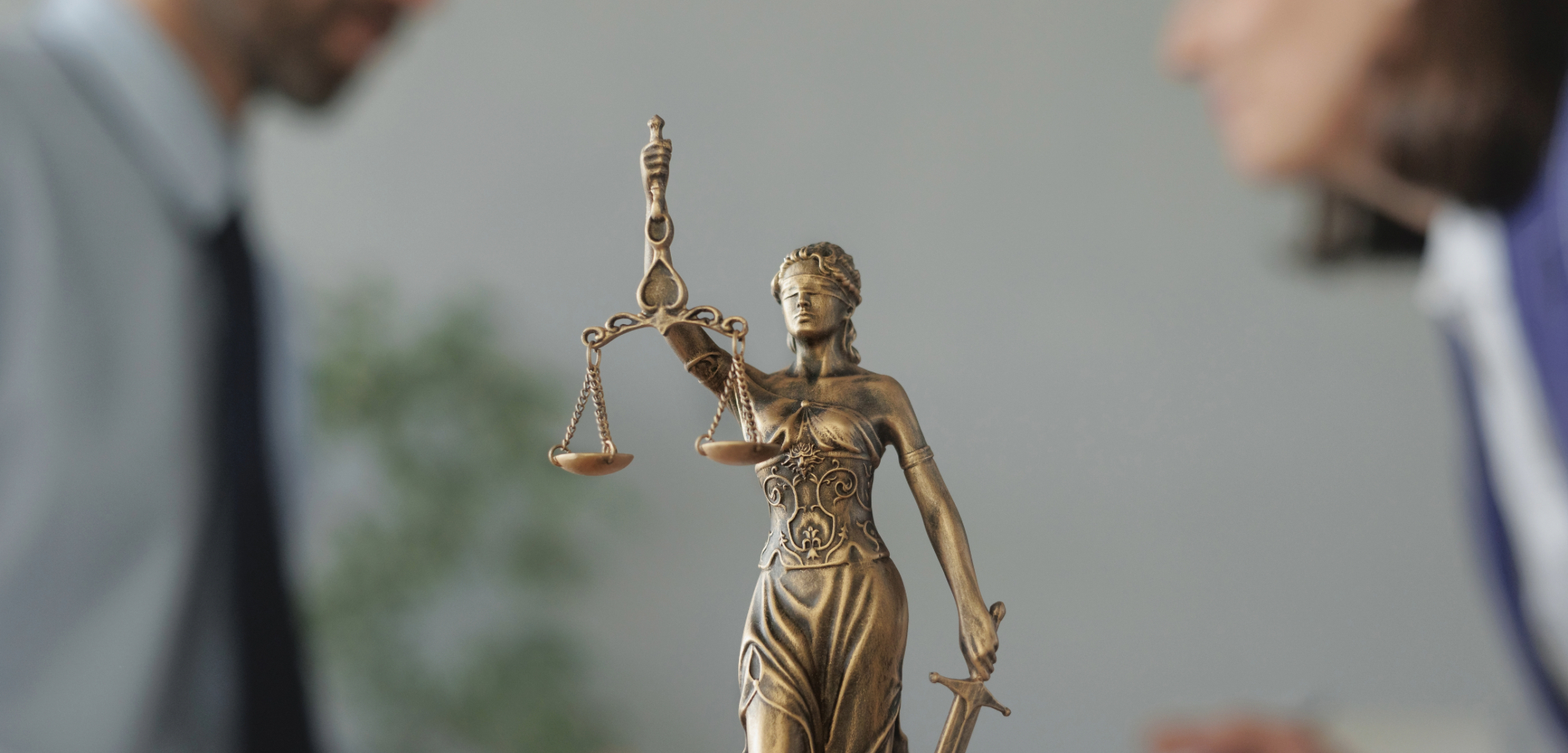 Estátua da deusa da justiça segurando balança em destaque, enquanto duas pessoas desfocadas conversam ao fundo, simbolizando imparcialidade e equidade no direito.