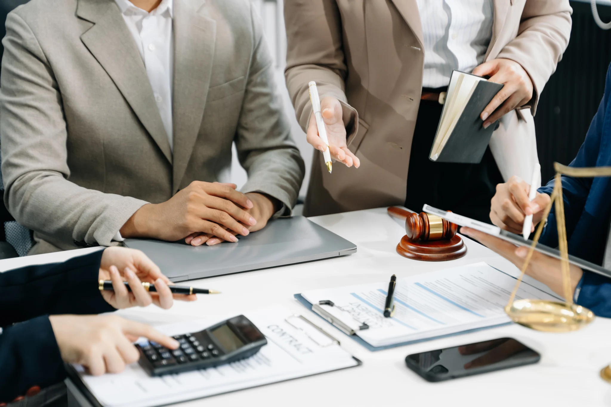 Equipe de advogados debate cláusulas de contrato sobre mesa branca, com martelo de juiz, balança da justiça, calculadora e laptop em destaque.
