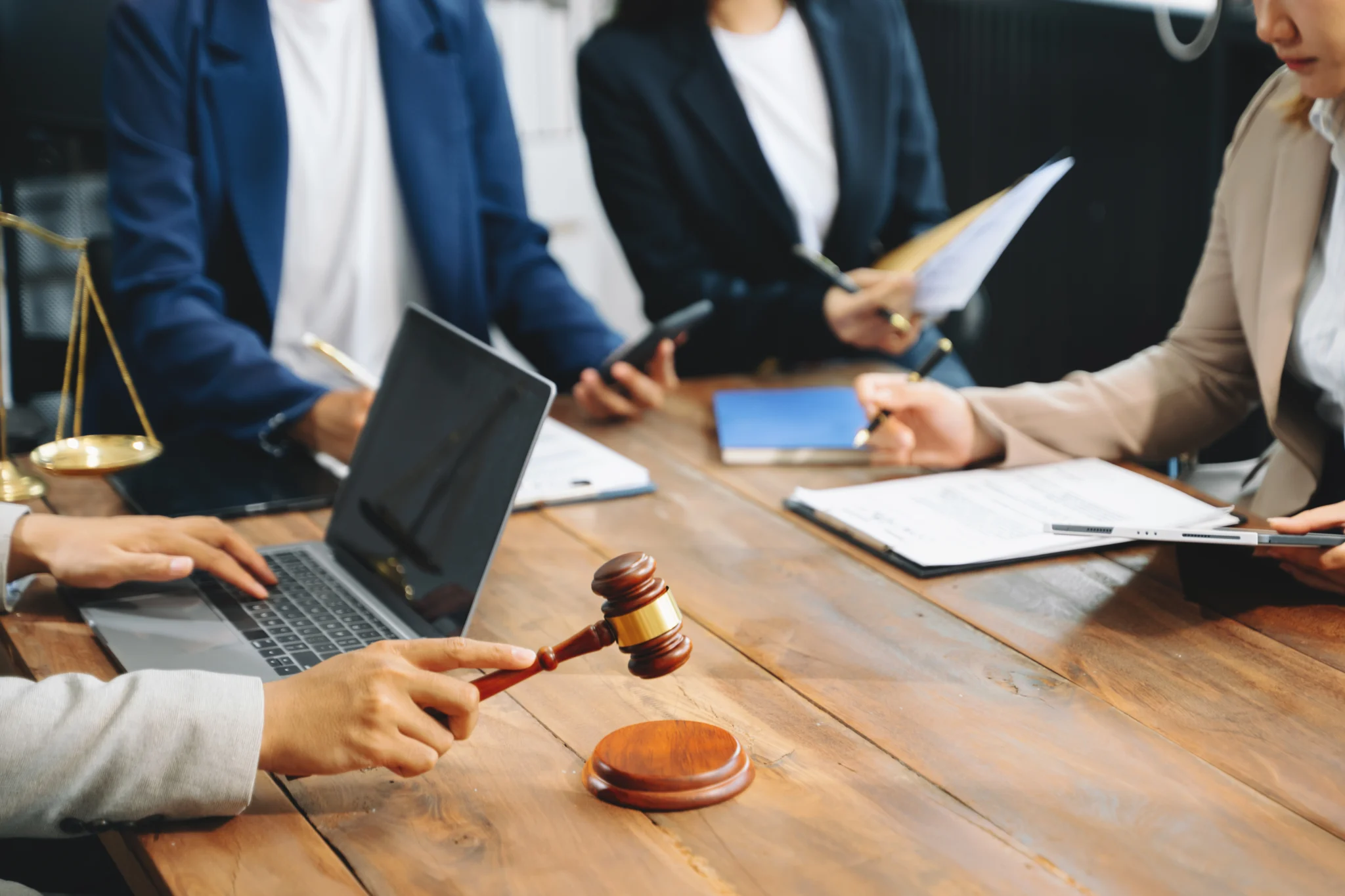 Advogados discutem caso em torno de mesa de madeira; um segura martelo de juiz, enquanto laptop, balança da justiça e documentos completam o cenário jurídico colaborativo.