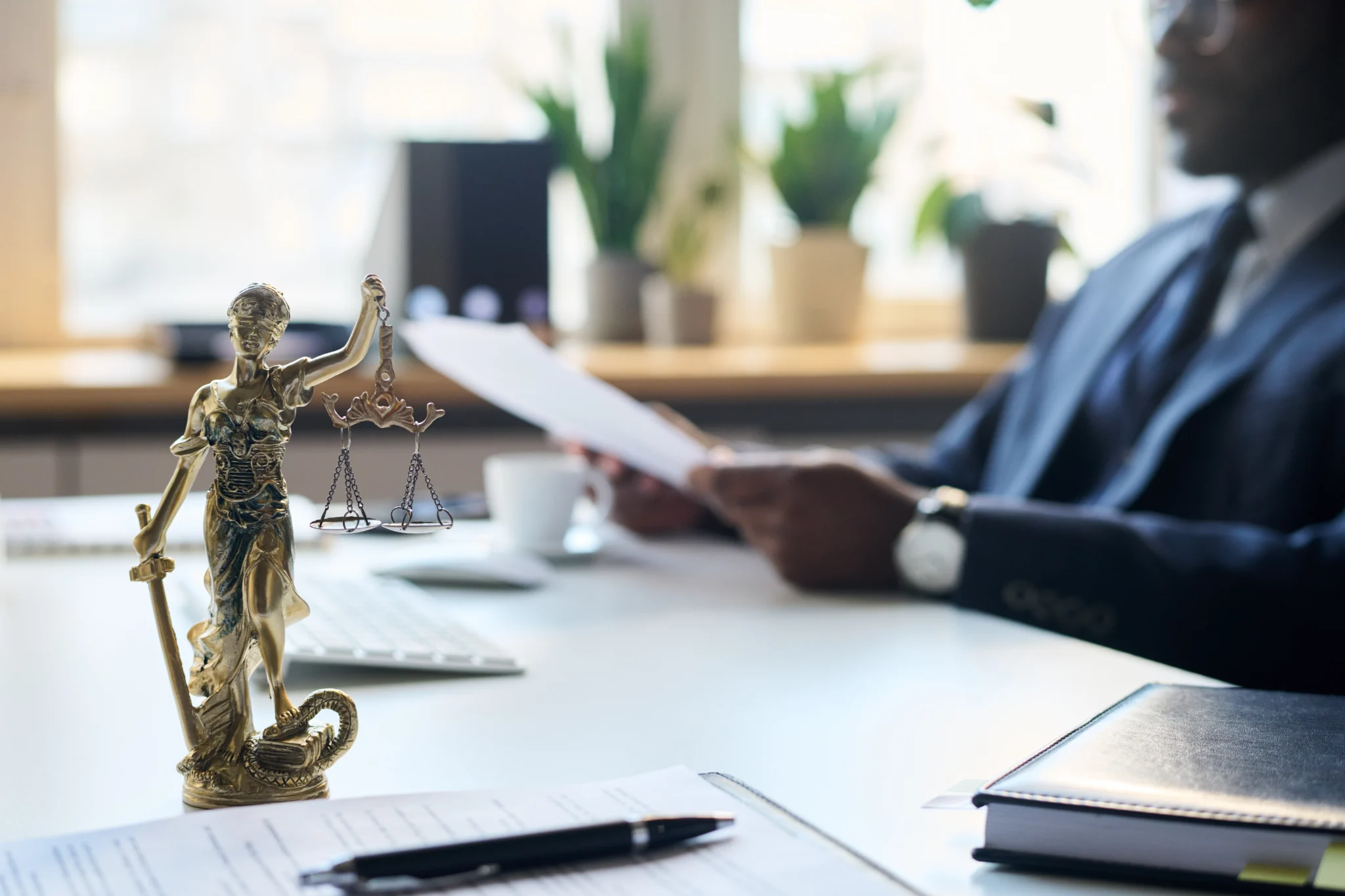 Estátua da deusa da justiça sobre a mesa em primeiro plano, enquanto advogado de terno lê documentos ao fundo, simbolizando equilíbrio e análise jurídica.
